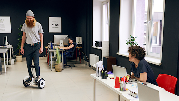 En person står på en eldriven balanscykel i ett kontorslandskap, medan två andra personer arbetar vid sina skrivbord.